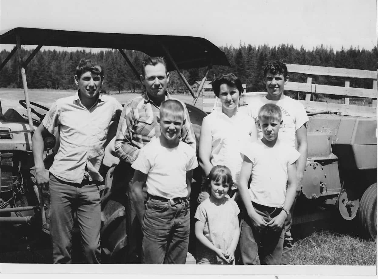 black and white photo of a family in jeans and a white shirt standing in front of a tractor in hay field