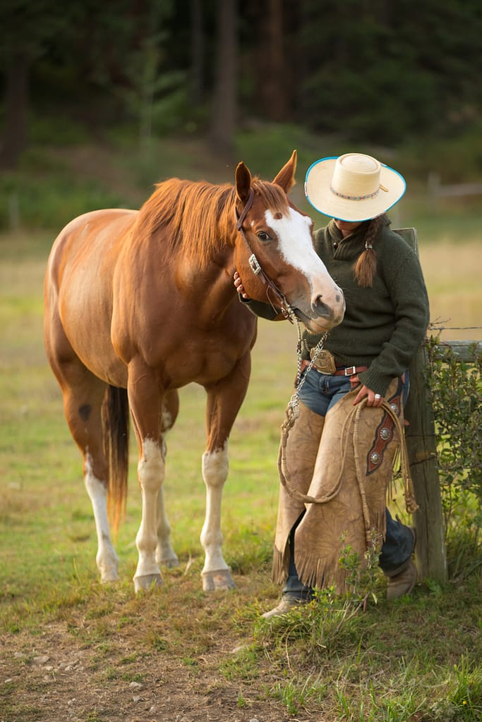 Cowgirl wearing a hat with sorrel horse by a fence