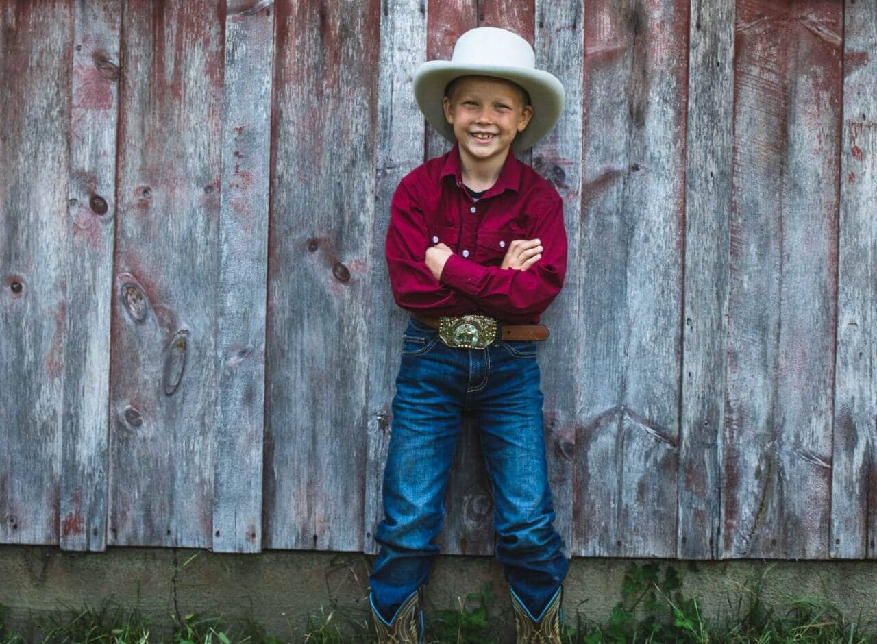 little boy in a cowboy hat standing in front of a faded red wall