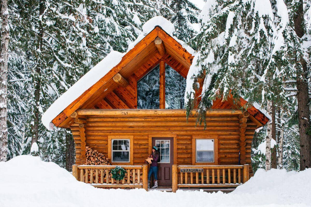 log cabin covered in snow covered forest with woman on the deck holding firewood