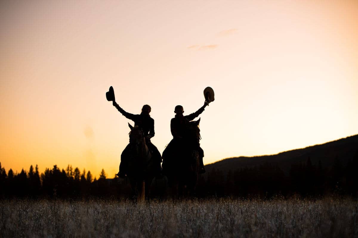 silhouette of cowboys on horseback with hats in the air and sunset behind them