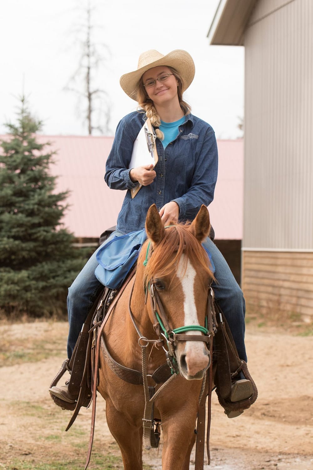 Cowgirl wearing glasses and holding a clipboard smiling and riding a dun Quarter Horse Mare